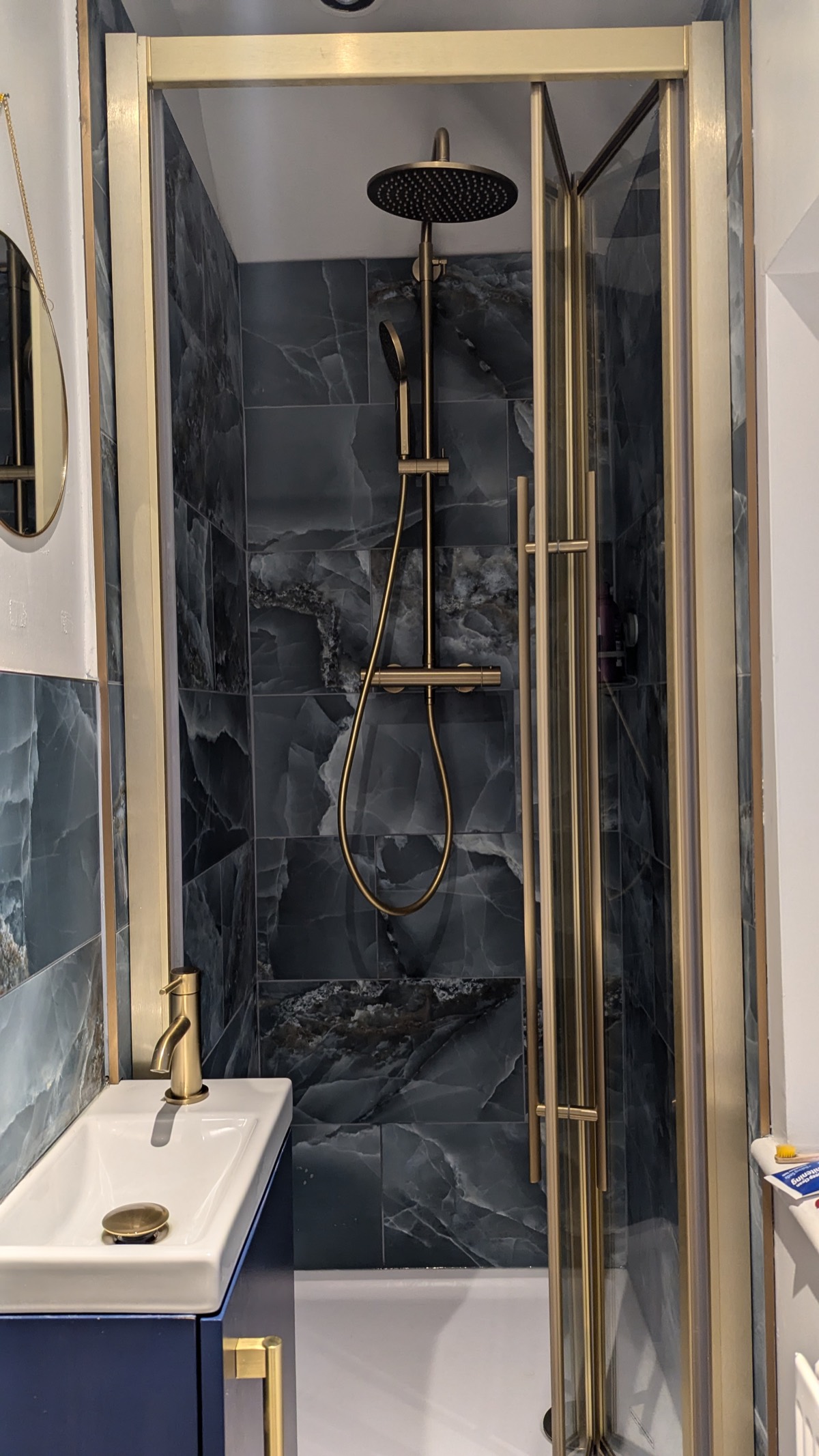 A brushed-brass framed shower enclosure over dark marble tiling, paired with a compact navy vanity and brass tap.